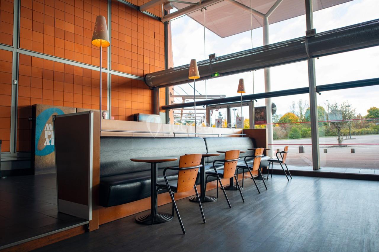 Intérieur d'un café moderne avec des tables, des chaises en bois et de grandes baies vitrées donnant sur l'extérieur.