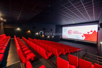 Salle de cinéma vide avec fauteuils rouges, grand écran allumé et plafond illuminé par des motifs lumineux.