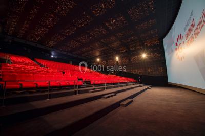 Salle de cinéma vide avec fauteuils rouges, grand écran allumé et plafond illuminé par des motifs lumineux.