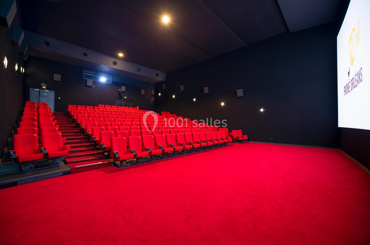 Salle de cinéma vide avec des rangées de fauteuils rouges, un écran blanc allumé et un sol recouvert de moquette rouge.