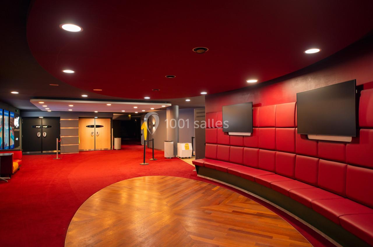 Hall d'entrée moderne avec sièges rouges, écrans muraux et sol en bois, éclairé par des spots au plafond.