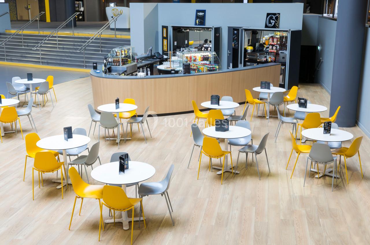 Espace cafétéria moderne avec tables rondes blanches, chaises jaunes et grises, et un comptoir de restauration en arrière…