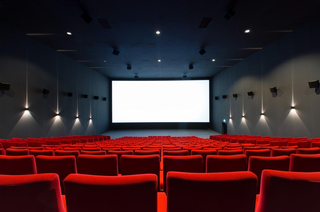 Salle de cinéma vide avec des rangées de sièges rouges et un grand écran blanc allumé.