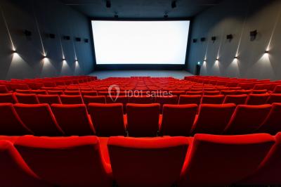 Salle de cinéma vide avec des rangées de sièges rouges et un grand écran blanc allumé.