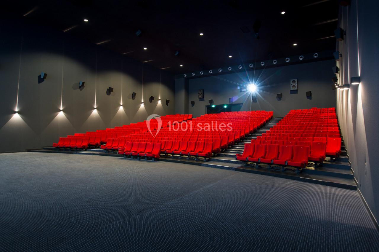 Salle de cinéma vide avec sièges rouges alignés et écran éteint, éclairée par des lumières murales et un projecteur.