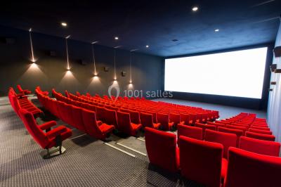 Salle de cinéma vide avec des rangées de sièges rouges et un grand écran blanc allumé.