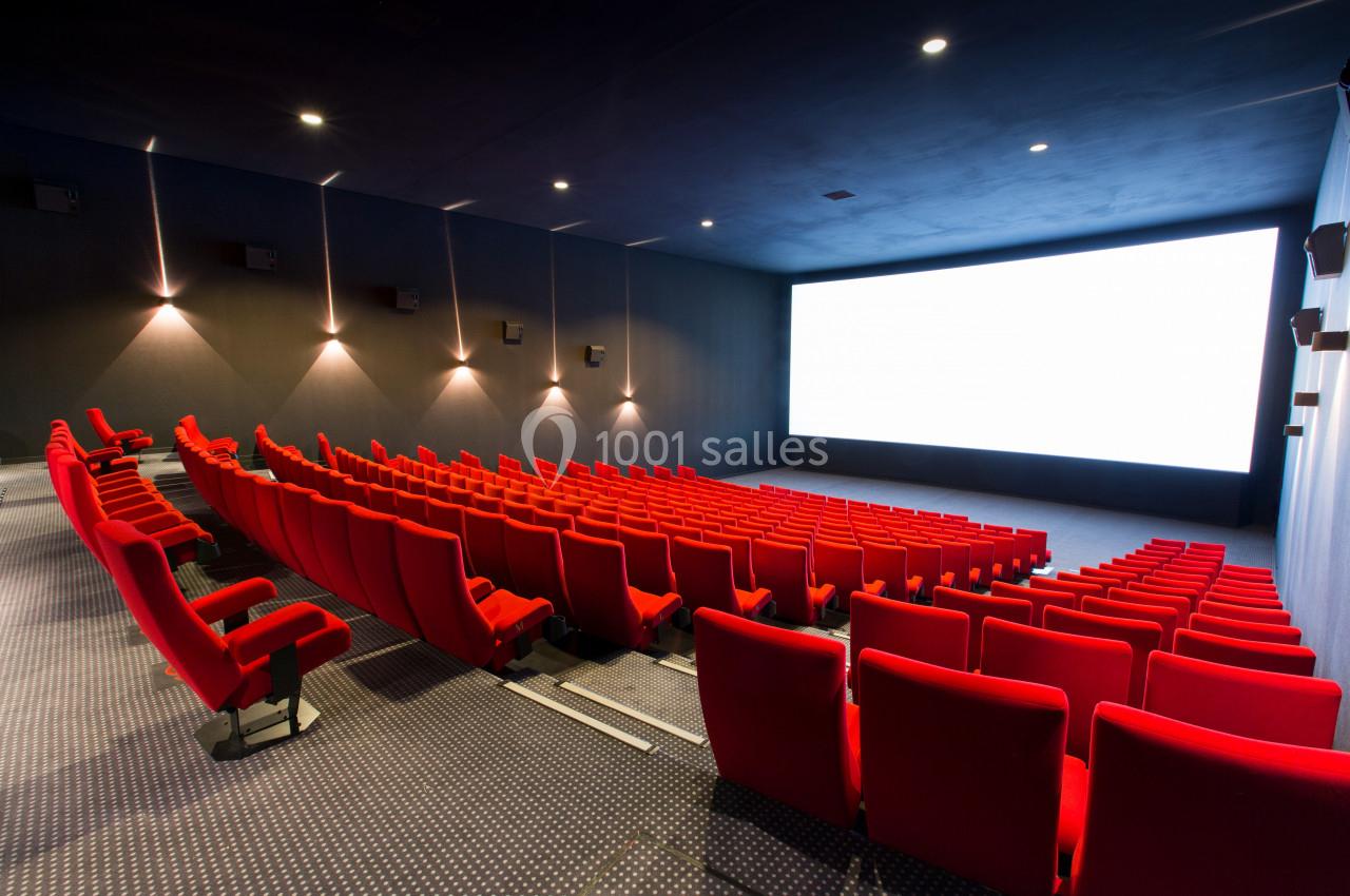 Salle de cinéma vide avec des rangées de fauteuils rouges et un grand écran blanc illuminé.