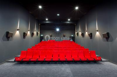 Salle de cinéma vide avec des rangées de sièges rouges et un grand écran blanc allumé.