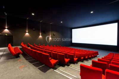 Salle de cinéma vide avec des rangées de sièges rouges et un grand écran blanc allumé.