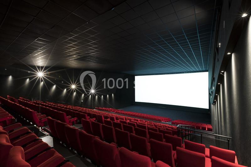 Salle de cinéma vide avec des rangées de sièges rouges et un grand écran blanc allumé.