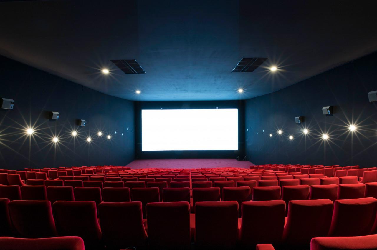 Salle de cinéma vide avec des rangées de sièges rouges et un grand écran blanc allumé au fond.