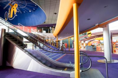 Escalier mécanique dans un espace intérieur moderne avec moquette violette, plafonds hauts et enseignes lumineuses.
