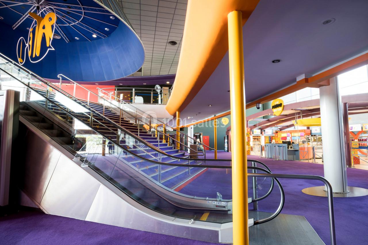 Escalier mécanique dans un espace intérieur moderne avec moquette violette, plafonds hauts et enseignes lumineuses.