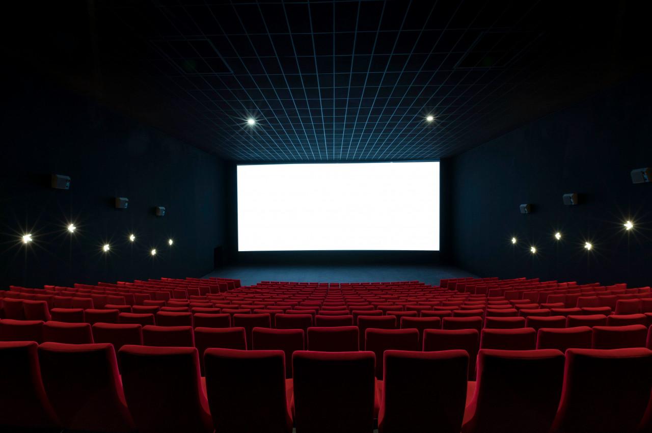 Salle de cinéma vide avec rangées de fauteuils rouges et un grand écran blanc allumé.