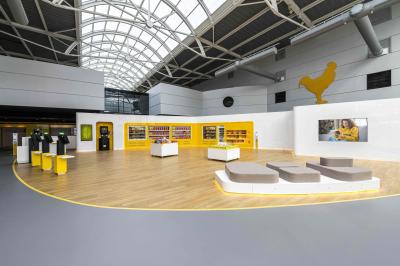 Couloir lumineux avec moquette grise, fauteuils, murs sombres, panneaux jaunes et ascenseurs numérotés 10.