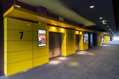 Couloir lumineux avec moquette grise, fauteuils, murs sombres, panneaux jaunes et ascenseurs numérotés 10.