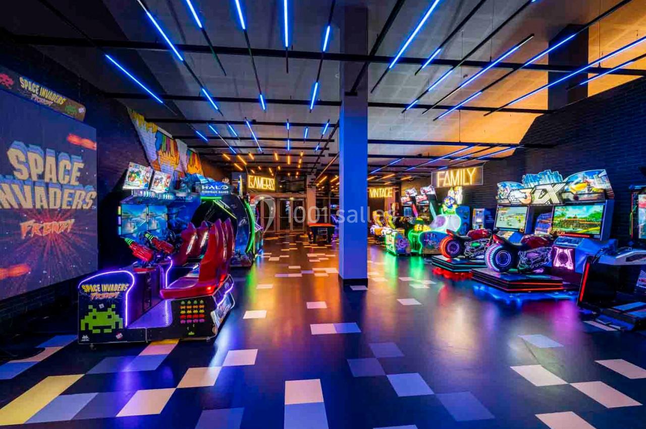 Salle d'arcade colorée avec des jeux vidéo rétro et modernes, éclairée par des néons bleus et multicolores.