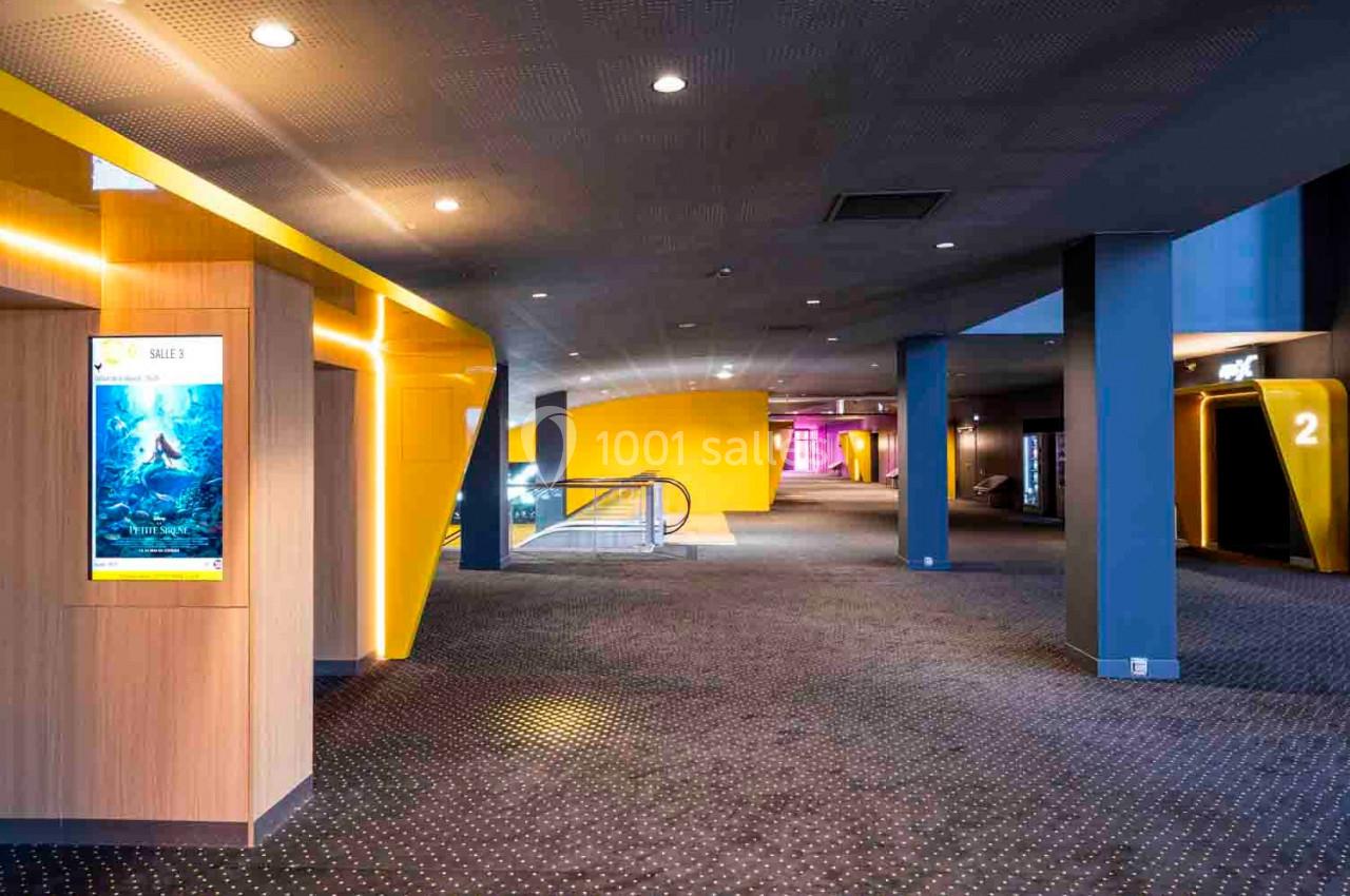 Hall d'entrée d'un cinéma moderne avec moquette sombre, murs jaunes et panneaux indiquant les salles.