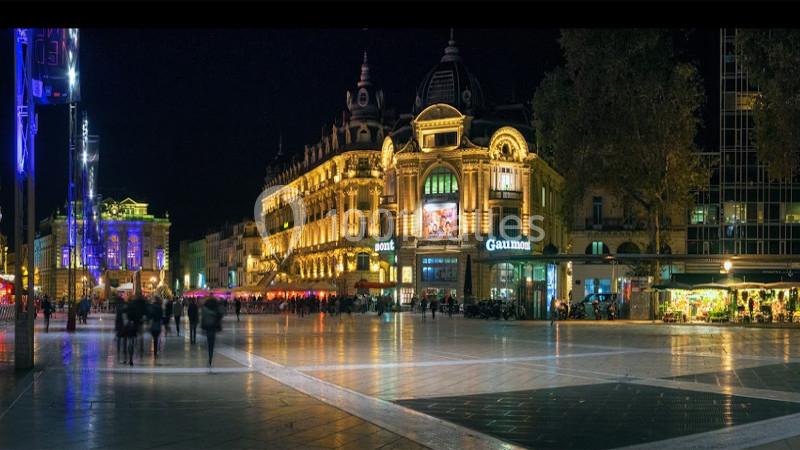 Place animée de nuit avec bâtiments illuminés, boutiques, et passants sur une large esplanade.