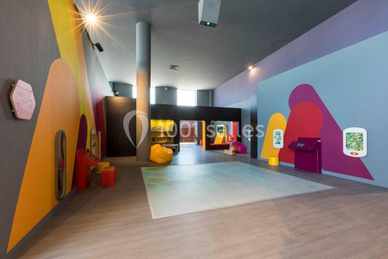 Salle interactive colorée avec des bornes numériques, des sièges et un espace au sol pour des activités.