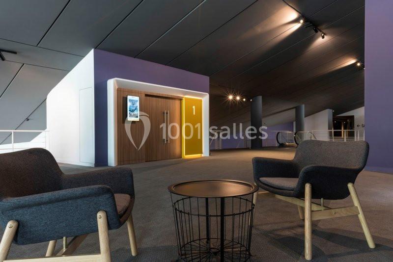 Deux fauteuils et une table basse dans un espace moderne avec un ascenseur encadré de lumière jaune au fond.