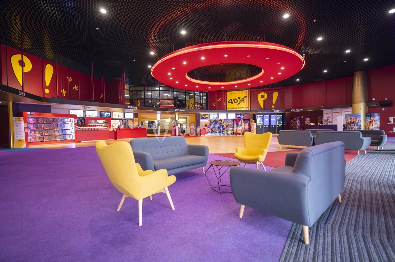 Hall de cinéma moderne avec fauteuils colorés, comptoirs de billetterie et éclairage circulaire rouge au plafond.