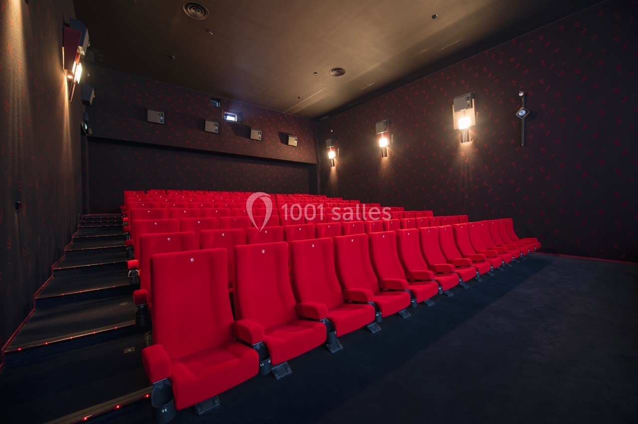 Salle de cinéma vide avec des rangées de fauteuils rouges et des murs sombres éclairés par des appliques murales.