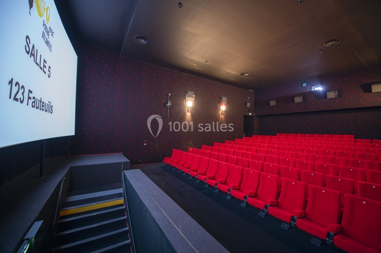 Salle de cinéma vide avec des rangées de fauteuils rouges, écran blanc et éclairage tamisé.