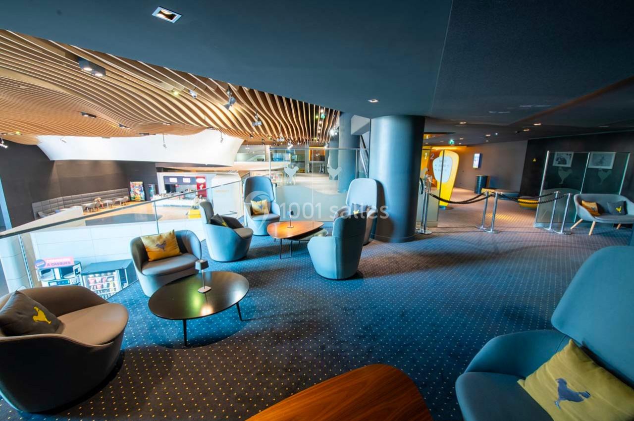 Espace lounge moderne avec fauteuils, tables basses et vue sur un hall lumineux, dans un décor aux tons bleus et bois.