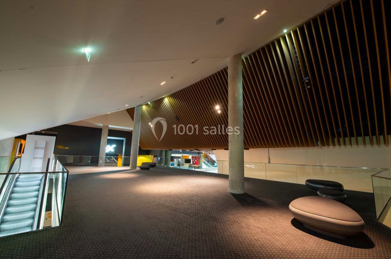 Intérieur moderne avec escalator, plafond incliné en bois, colonnes et sièges arrondis sur un sol sombre.