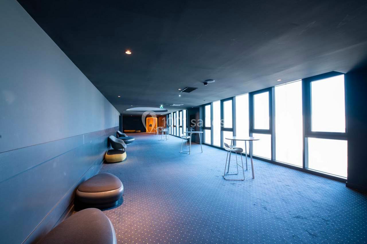 Couloir spacieux avec moquette bleue, grandes fenêtres laissant entrer la lumière, chaises et tables dispersées.