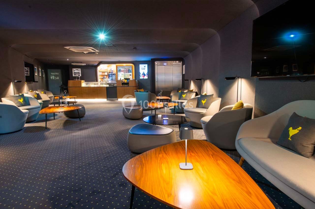 Salon moderne avec fauteuils gris, tables en bois, éclairage tamisé et un bar en arrière-plan.