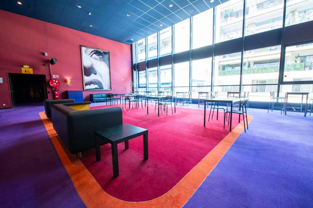 Salle lumineuse avec de grandes baies vitrées, tables et chaises modernes, décorée de couleurs vives et d'un tableau mural.
