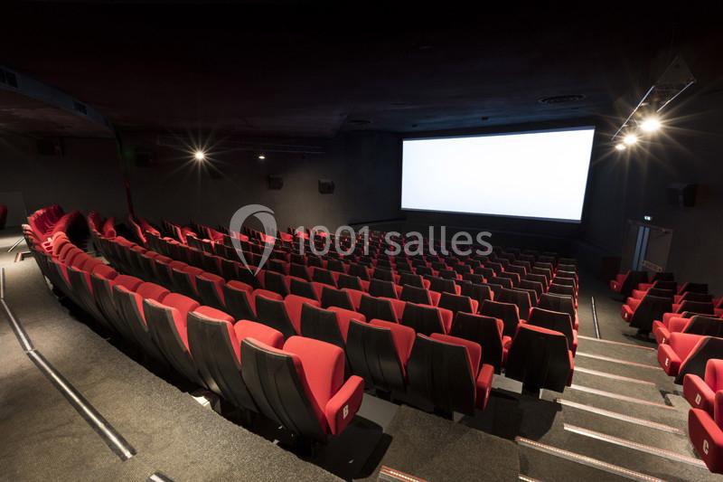 Salle de cinéma vide avec rangées de sièges rouges et écran blanc allumé dans un environnement sombre.