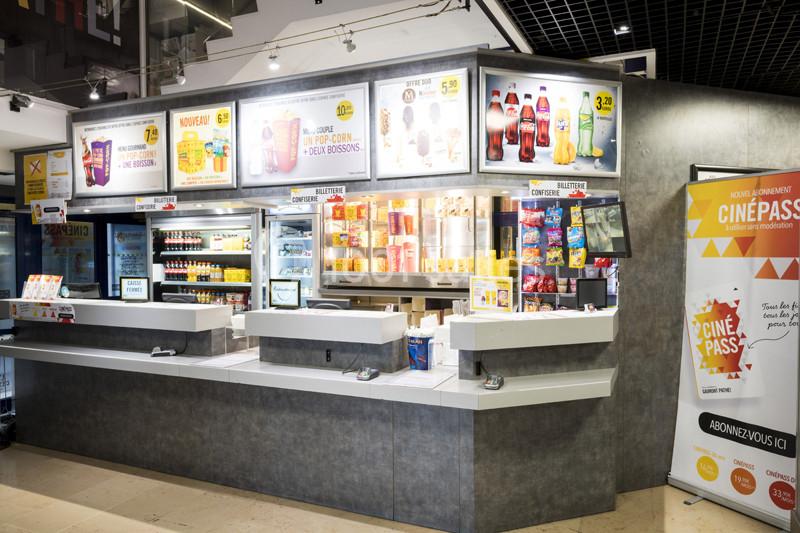 Comptoir de vente dans un cinéma proposant des boissons, snacks et menus affichés sur des panneaux lumineux.