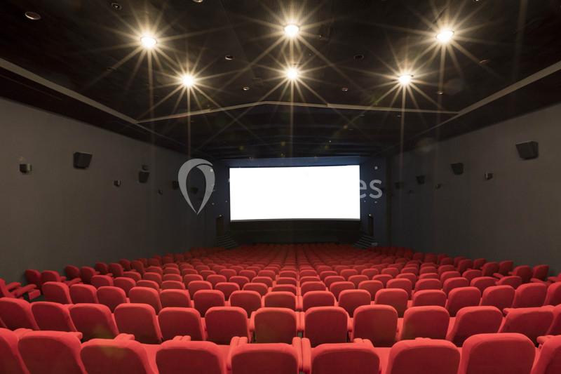 Salle de cinéma vide avec des rangées de sièges rouges et un écran blanc allumé au fond.
