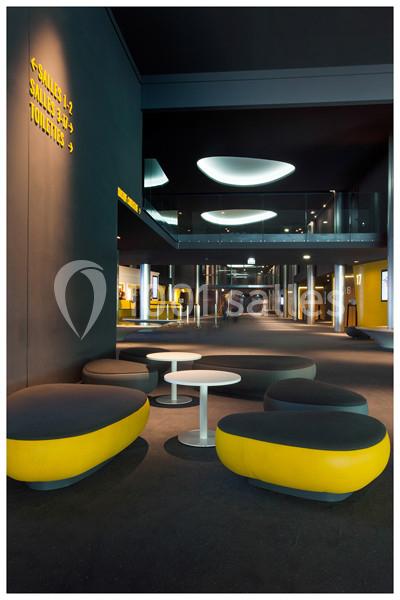 Espace intérieur moderne avec sièges jaunes et noirs, tables basses blanches et signalétique murale indiquant des salles.