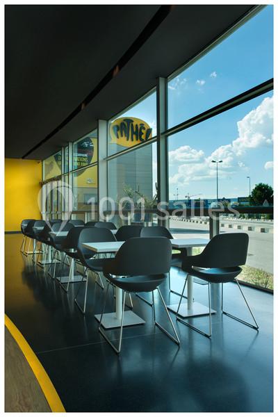 Rangée de tables et chaises modernes près de grandes fenêtres donnant sur un parking et un ciel dégagé.