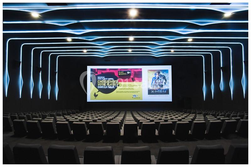 Salle de cinéma moderne avec rangées de sièges vides et un grand écran affichant des publicités et une affiche de film.