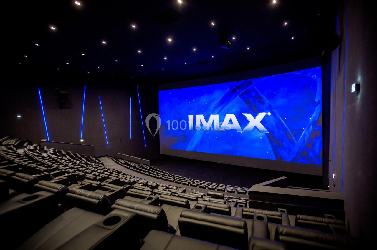 Salle de cinéma IMAX vide avec sièges en gradins, grand écran allumé affichant le logo IMAX et éclairage tamisé.