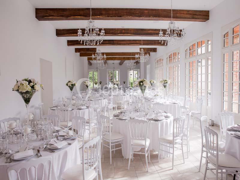 Salle de réception lumineuse avec tables rondes dressées, chaises blanches, décorations florales et lustres suspendus.