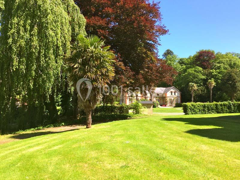 Grande maison entourée d'arbres, pelouse bien entretenue et ciel bleu en arrière-plan.