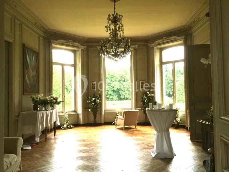 Salon lumineux avec parquet en bois, grandes fenêtres donnant sur un jardin, mobilier élégant et lustre central.