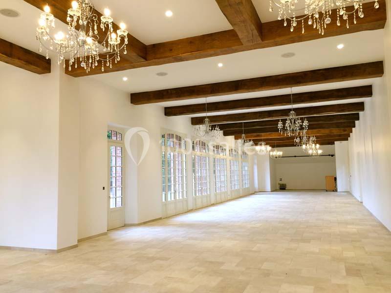 Grande salle lumineuse avec poutres en bois, lustres suspendus et larges fenêtres donnant sur l'extérieur.