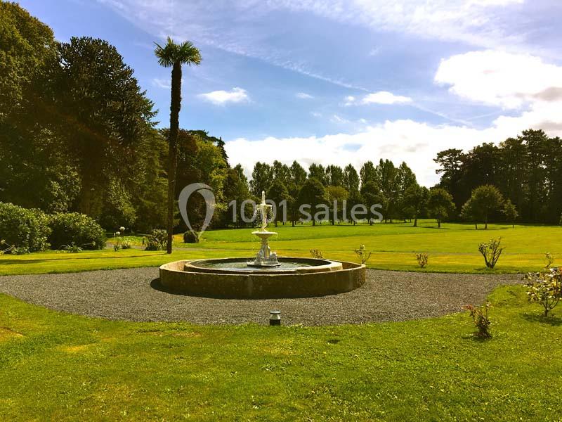 Fontaine centrale entourée de pelouse et d'arbres dans un parc sous un ciel ensoleillé.