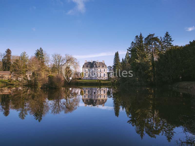 Château entouré d'arbres, reflété dans un lac calme sous un ciel bleu clair.