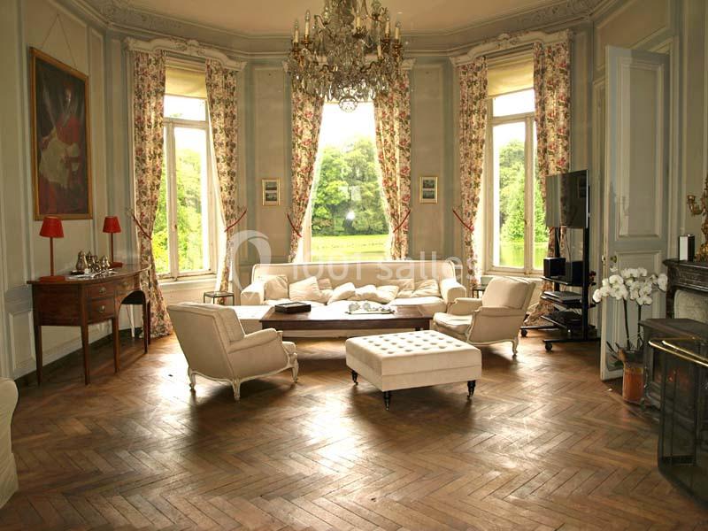 Salon élégant avec parquet en chevrons, mobilier classique, chandelier et grandes fenêtres donnant sur un jardin verdoyant.