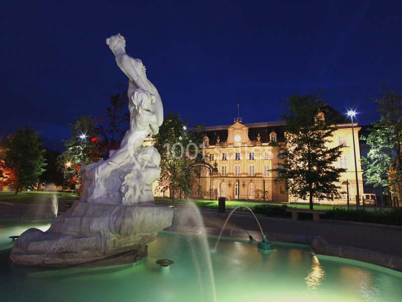 Statue éclairée dans une fontaine au premier plan, avec un bâtiment historique illuminé en arrière-plan, de nuit.