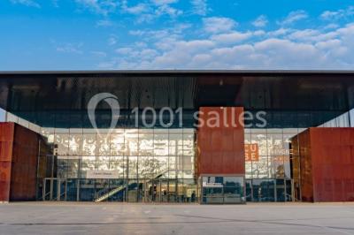 Façade moderne d'un bâtiment avec des murs en verre et acier rouillé, sous un ciel partiellement nuageux.