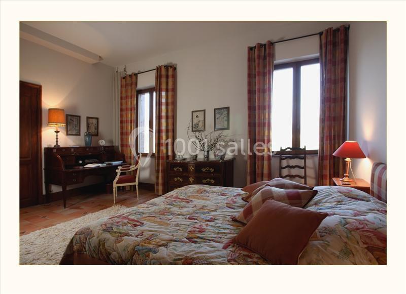 Chambre lumineuse avec lit double, bureau en bois, lampes allumées et rideaux à carreaux devant des fenêtres.